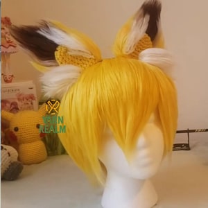 May include: A yellow wig with fox ears, featuring a blend of yellow, white, and brown. The ears are attached to a yellow headband. The wig is styled on a white mannequin head. The logo "YARN REALM" is visible.