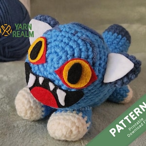 May include: A handmade blue crochet monster with large yellow and black eyes, white teeth, and white paws. The monster has white ear accents and is labelled "PATTERN Printable Download File". The Yarn Realm logo is visible in the upper left corner.