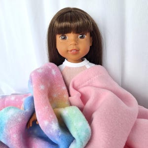 Puede incluir: Una muñeca con cabello castaño y pecas está envuelta en una manta suave y colorida. La manta presenta un diseño de arcoíris pastel con acentos blancos en forma de estrella. Una manta rosa sólida también está envuelta alrededor de la muñeca.