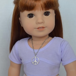 Könnte beinhalten: Eine Puppe mit langen, glatten, rotbraunen Haaren, die ein lavendelfarbenes T-Shirt und eine Halskette mit einem Friedenssymbol trägt. Die Puppe trägt einen weißen und rosa gemusterten Rock.