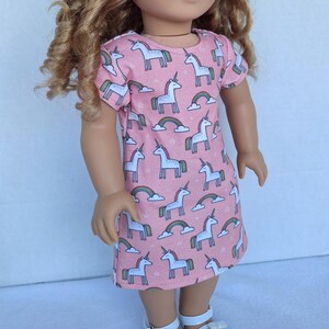 Könnte beinhalten: Ein rosa Puppenkleid mit einem Muster aus weißen Einhörnern, Regenbogen und Wolken. Das kurzärmelige Kleid ist für eine Puppe entworfen und hat einen runden Ausschnitt. Das Einhorn-Design wiederholt sich auf dem Stoff.