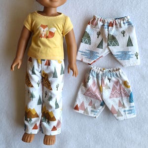 Può includere: Una bambola indossa una maglietta gialla con una grafica di volpe e pantaloni bianchi con un motivo di montagne e alberi. Sono mostrati anche due paia di pantaloncini abbinati, con la stessa stampa. L'abbigliamento è progettato per una bambola.
