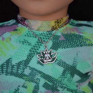 Op de afbeelding: Een zilveren ketting met een lotusbloem hanger met een persoon die in meditatieve houding zit. De ketting wordt gedragen door een pop met een groen, roze en geel gepatroneerd shirt.
