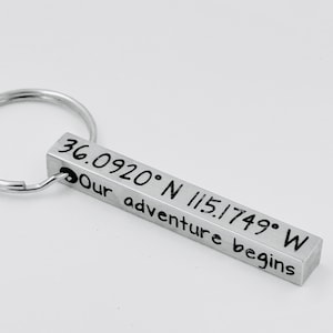 May include: A silver metal keychain with a rectangular pendant. The pendant is engraved with coordinates and the phrase "Our adventure begins". The coordinates are 36.0920° N 115.1749° W.