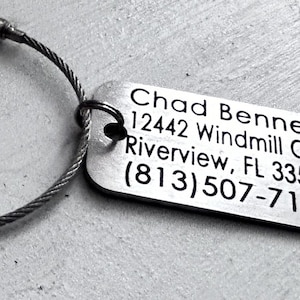 May include: Silver metal keychain with a cable chain and a rectangular tag engraved with the text "Chad Bennett, 12442 Windmill Cove Dr, Riverview, FL 33569, (813) 507-7137".