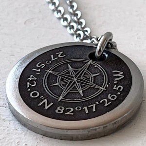 May include: A silver necklace with a round pendant featuring a compass design and the coordinates "0° 42' 51.5" N 82° 17' 26.5" W".