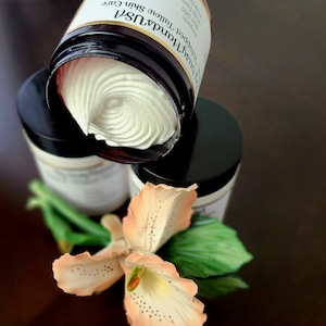 May include: A close-up of a jar of organic, whipped tallow skin care with the label "Handcrafted USA" and "Tallow Skin Care" printed on it. The jar is open and the cream is visible. The jar is sitting on a brown surface with a flower.