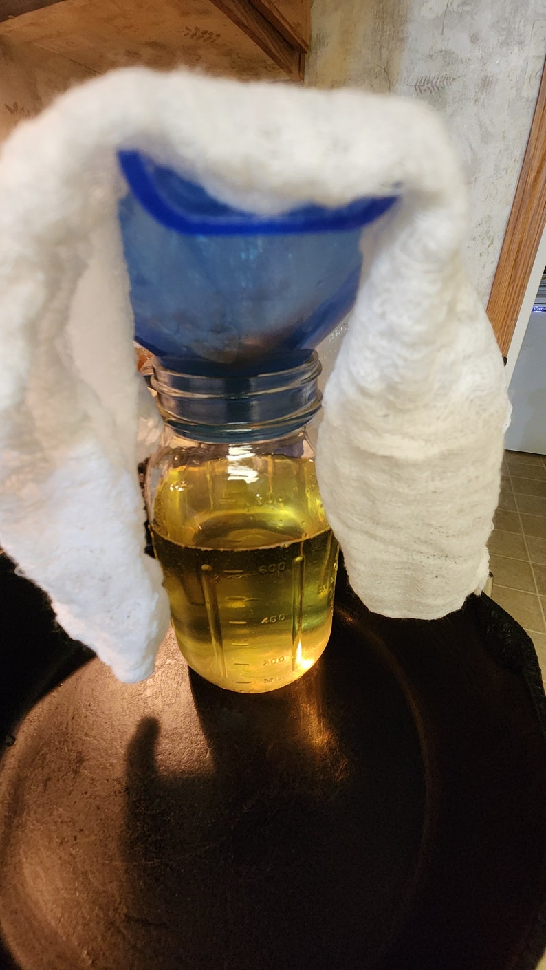 Pu&ograve; includere: Un barattolo di vetro trasparente con un liquido giallo al suo interno, appoggiato su una superficie nera. Un imbuto blu &egrave; posizionato sopra il barattolo, e un panno bianco &egrave; drappeggiato sopra l'imbuto.