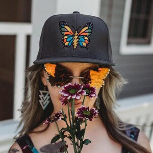 Può includere: Un cappellino da baseball grigio scuro con una farfalla ricamata dalle ali arancioni, blu e gialle e il corpo a forma di fulmine. È abbinato a occhiali da sole a forma di farfalla con lenti arancioni e orecchini triangolari color argento.