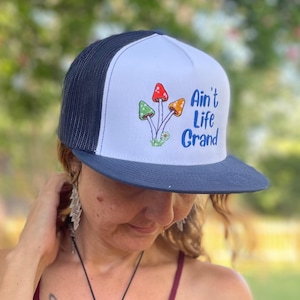 May include: A white and dark blue trucker hat featuring a graphic of three mushrooms in red, green, and orange with the text Ain't Life Grand in blue script. The hat has a solid white front panel, a dark blue brim, and dark blue mesh back panels.