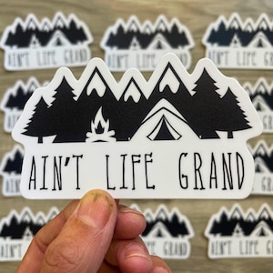 May include: White sticker with a black mountain range, trees, campfire, and tent design. The text "AIN'T LIFE GRAND" is printed below the graphic. The sticker is rectangular with a white border and is suitable for outdoor use.