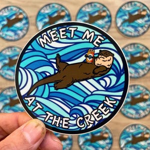 May include: A round sticker featuring a cartoon otter floating in blue water, holding a can of beer. The text "MEET ME AT THE CREEK" is in a white, bold font around the otter. Additional stickers with the same design are in the background.