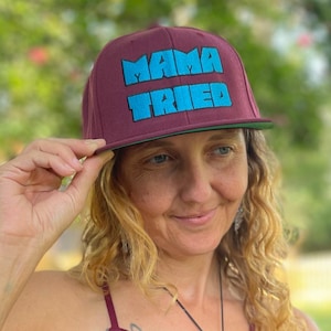 May include: A maroon baseball cap with a flat brim and the text MAMA TRIED embroidered in bright blue block letters across the front panel. The cap features a structured crown and a green underside on the brim, providing a bold contrast.
