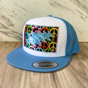 May include: A light blue trucker hat with a white front panel and a blue mesh back. The front features a patch with a tie-dye lightning bolt design over a background of colorful peace signs. The hat has a curved brim.