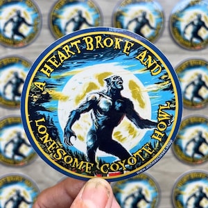 Op de afbeelding: Ronde sticker met een blauwe rand en de tekst "A HEART BROKE AND LONESOME COYOTE HOWL". De sticker toont een weerwolf die naar een volle maan huilt. De achtergrond is blauw en geel. De sticker wordt door een hand vastgehouden.