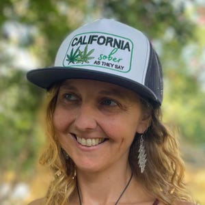 May include: A white and black trucker cap featuring a green graphic of two cannabis leaves and the text CALIFORNIA sober AS THEY SAY printed in black and green on the front panel. The cap has a solid white front and a black mesh back.