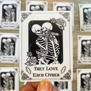 May include: White rectangular sticker featuring a black and white illustration of two skeletons embracing. The text "THEY LOVE EACH OTHER" is below the illustration. Roses and decorative elements frame the image.