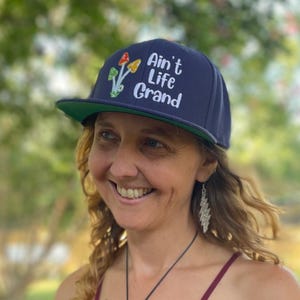 Puede incluir: Una gorra de béisbol azul marino con visera plana y parte inferior verde. La parte frontal presenta un diseño bordado de tres setas coloridas y el texto blanco "Ain't Life Grand" en una fuente redondeada y divertida.