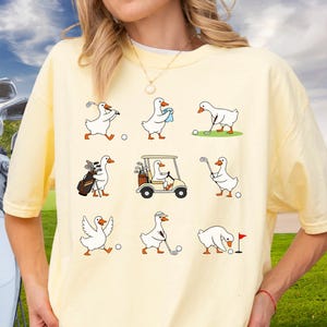 Peut inclure: T-shirt jaune clair avec un motif répété d'oies de dessin animé jouant au golf. Les oies sont représentées avec des clubs de golf, des voiturettes de golf et des balles de golf. Le t-shirt est de style décontracté.