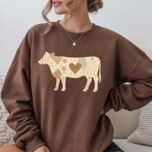 Può includere: Una felpa marrone con un design a patchwork di mucca. L'illustrazione della mucca incorpora vari motivi, tra cui floreali, a quadri e a forma di cuore, nelle tonalità del beige, marrone e crema. La felpa è di colore marrone tinta unita.