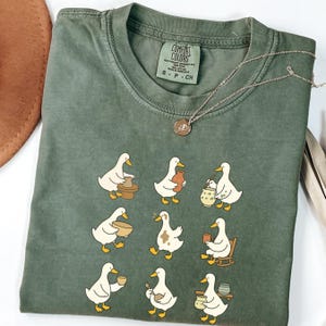 Puede incluir: Camiseta verde oliva Comfort Colors con un diseño caprichoso de patos creando cerámica. Los patos se representan en varias poses, trabajando con ollas de arcilla y otras herramientas de cerámica. Un collar plateado con un pequeño colgante descansa sobre la camiseta.