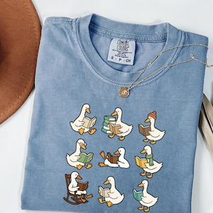 May include: A pale blue Comfort Colors t-shirt with a design of cartoon ducks reading books. The ducks are in different poses, some with accessories. A gold necklace with a pendant is draped across the shirt.