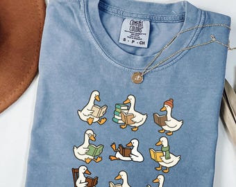 Puede incluir: Una camiseta Comfort Colors azul claro con un diseño de patos de dibujos animados leyendo libros. Los patos están en varias poses, algunos con accesorios. Un collar dorado con un colgante está sobre la camiseta.