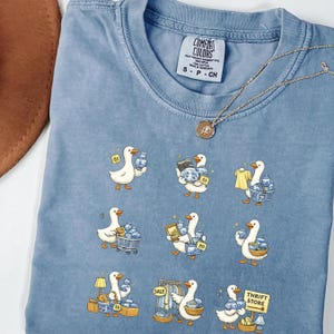 以下が含まれることがあります： 水色のComfort Colors Tシャツ。青と白の陶器を持ったアヒルのグラフィックが特徴です。クルーネックと半袖です。ペンダント付きのゴールドネックレスがシャツの上にかけられています。
