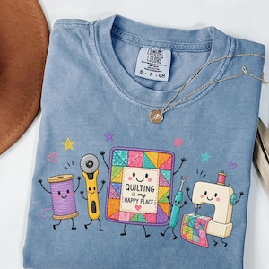 以下が含まれることがあります： 青い半袖Tシャツ。紫色の糸巻き、黄色のロータリーカッター、"QUILTING is my HAPPY PLACE"という文字が入ったカラフルなキルトブロック、ミシンなど、裁縫道具をモチーフにした漫画風のグラフィックプリントが施されています。