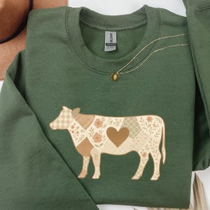 Puede incluir: Sudadera verde oscuro con un diseño de vaca en un patrón de patchwork en tonos marrón y crema, con un detalle de corazón. Un collar dorado con un colgante está sobre la sudadera.