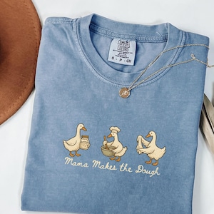 Puede incluir: Una camiseta azul Comfort Colors con un gráfico de tres patos de dibujos animados, cada uno involucrado en una actividad de horneado. El texto "Mama Makes the Dough" está debajo. Un collar de oro con un colgante está sobre la camiseta.