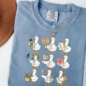 Könnte beinhalten: Ein blaues Kurzarm-T-Shirt mit einem Grafikdruck von neun weißen Enten, die als Camp-Betreuer gekleidet sind. Jede Ente hält verschiedene Gegenstände wie eine Wasserflasche, ein Schild, ein Klemmbrett, ein Megafon, eine Flagge oder einen Rucksack.