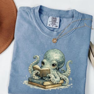 Puede incluir: Una camiseta azul claro con un gráfico de un pulpo verde claro leyendo un libro abierto mientras descansa sobre una pila de dos libros. La camiseta tiene una etiqueta de la marca Comfort Colors en el cuello y una fina cadena dorada sobre la tela.