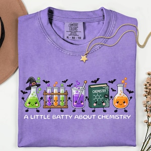 May include: A lavender t-shirt featuring a graphic of smiling chemistry equipment including beakers, test tubes, and a book. The text reads A LITTLE BATTY ABOUT CHEMISTRY, accompanied by small black bat illustrations and a gold star necklace.