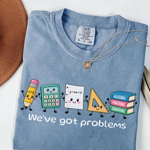 Puede incluir: Una camiseta azul de cuello redondo con un gráfico de un lápiz, calculadora, papel, regla triangular y una pila de libros de dibujos animados con el texto "We've got problems". La camiseta tiene una etiqueta de marca Comfort Colors en el cuello.