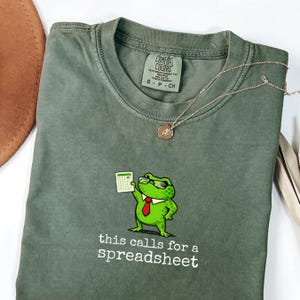 Può includere: T-shirt verde salvia con un ranocchio dei cartoni animati con occhiali e cravatta rossa, che tiene un foglio di calcolo. Il testo "this calls for a spreadsheet" è sotto. È visibile anche una collana d'argento con un piccolo ciondolo.