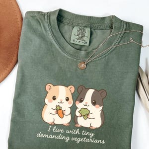 May include: Sage green t-shirt with a graphic of two cartoon hamsters, one holding a carrot and the other broccoli. The text "I live with tiny demanding vegetarians" is below the hamsters. A silver necklace with a pendant is on the shirt.