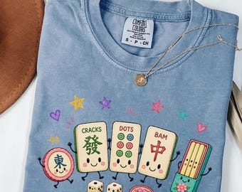 May include: A blue short-sleeve t-shirt featuring a graphic of smiling mahjong tiles and dice. The print includes the text CRACKS, DOTS, and BAM above the tiles. A gold-toned necklace with a small circular pendant rests on the neckline.