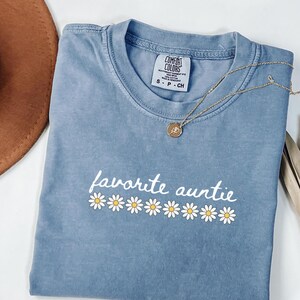Peut inclure: Un t-shirt bleu Comfort Colors avec l'inscription "favorite auntie" en écriture blanche et des marguerites. Un collier en or avec un pendentif est posé sur le t-shirt. Un chapeau marron est visible en haut à gauche.