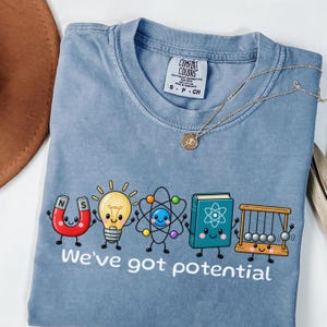 Peut inclure: Un t-shirt bleu à manches courtes avec un motif de dessin animé représentant un aimant, une ampoule, un atome, un livre et un pendule de Newton, accompagné du texte "We've got potential". Il présente un col rond et une étiquette Comfort Colors.