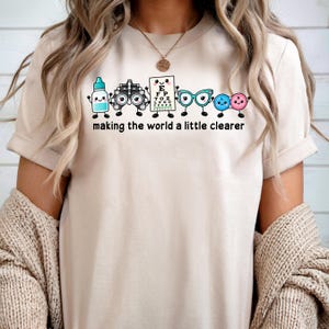 May include: A beige t-shirt featuring a graphic of cartoon eye care items including a bottle, phoropter, eye chart, glasses, and contact lenses with the text "making the world a little clearer" printed in black lettering below the characters.