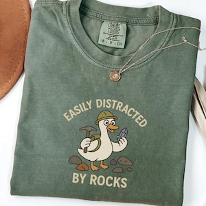 以下が含まれることがあります： 「EASILY DISTRACTED BY ROCKS」の文字と、鉱山用ヘルメット、つるはし、クリスタルを持った漫画のアヒルのイラストがプリントされたオリーブグリーンのTシャツ。ペンダント付きのゴールドネックレスがシャツの上にあります。