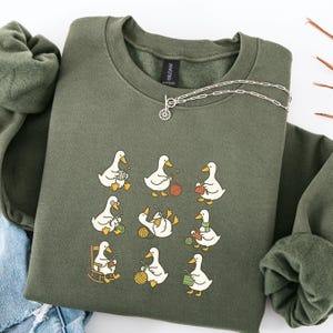 Puede incluir: Una sudadera verde oliva con un diseño caprichoso de patos blancos tejiendo con lana colorida. Los patos están en varias poses, algunos con gafas o bufandas. Un collar plateado con un colgante de sol está sobre la sudadera.