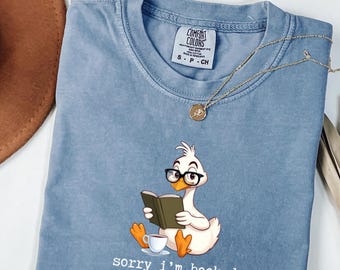 Puede incluir: Camiseta Comfort Colors azul claro con un pato de dibujos animados con gafas leyendo un libro, con una taza de café. El texto "sorry i'm booked" está debajo del pato. Un collar dorado está sobre la camiseta.
