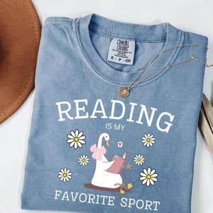 Op de afbeelding: Een blauw Comfort Colors t-shirt met de tekst "READING IS MY FAVORITE SPORT". Het shirt heeft een afbeelding van een gans die een boek leest, omringd door madeliefjes. Een gouden ketting met een hanger ligt over het shirt.