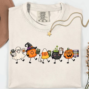 Può includere: Una t-shirt color crema con una grafica di cinque personaggi dei cartoni animati: un fantasma con gli occhiali, una zucca con un cappello da strega, un candy corn, un calderone ribollente e una zucca che tiene libri con scritto Invoices, Expenses, Payroll, Taxes.