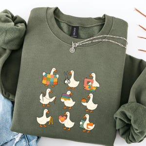 Pode incluir: Camisola verde-oliva com um design extravagante de gansos de desenho animado envolvidos em várias atividades de artesanato. Os gansos são retratados com máquinas de costura, tesouras e colchas. Um colar de prata com um pingente está drapeado sobre a camisola.