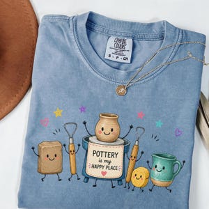 May include: A blue short-sleeved t-shirt with a graphic print featuring cartoon pottery tools and a pot with a face. The text reads POTTERY is my HAPPY PLACE. The shirt has a Comfort Colors label in the neck and a thin gold-tone chain necklace rests on top.