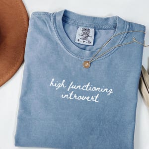 Könnte beinhalten: Ein blaues T-Shirt mit dem weißen Schriftzug "high functioning introvert". Eine goldene Halskette mit Anhänger liegt auf dem Shirt. Das Shirt ist gefaltet, und ein brauner Hut ist im Hintergrund zu sehen.