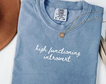 May include: A blue t-shirt with the words "high functioning introvert" in white script. A gold necklace with a pendant rests on the shirt. The shirt is folded, and a brown hat is visible in the background.